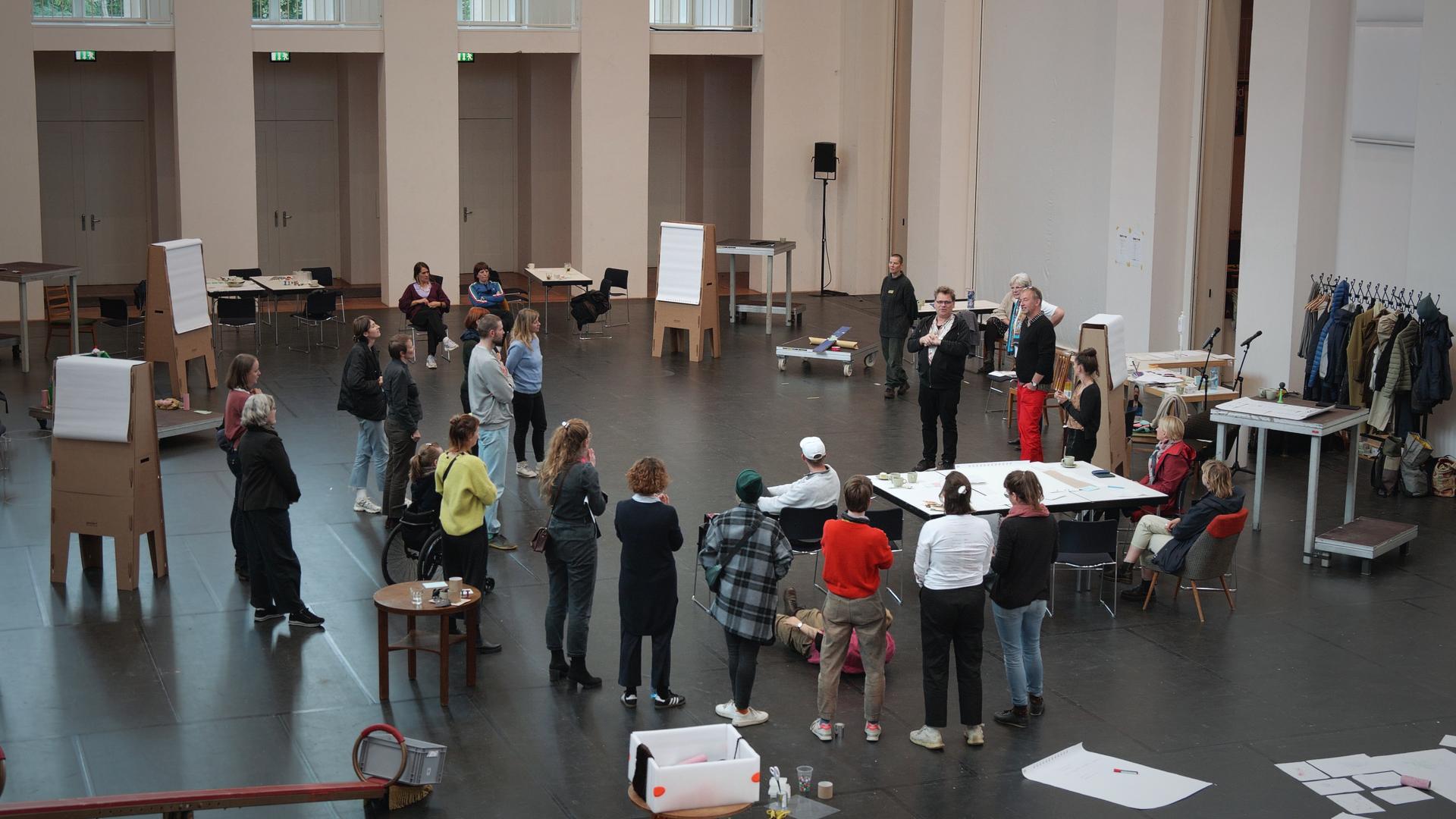 Eine Gruppe von Menschen im Kreis in einem Saal, umgeben von Flipcharts und Tischen
