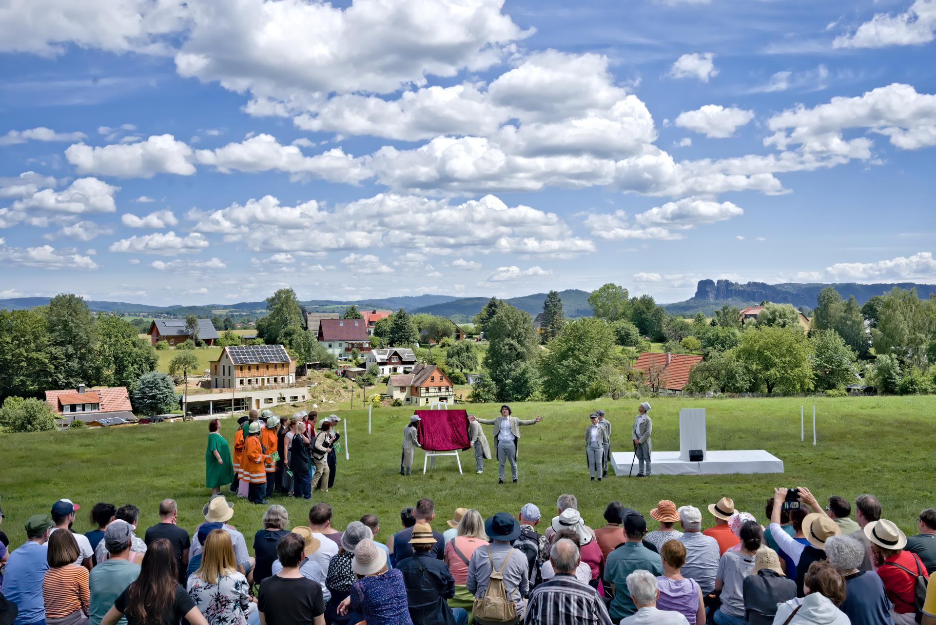 Eine Gruppe von Schauspieler*innen in Aktion, draußen auf der Wiese vor ländlicher Szenerie mit großem Publikum 