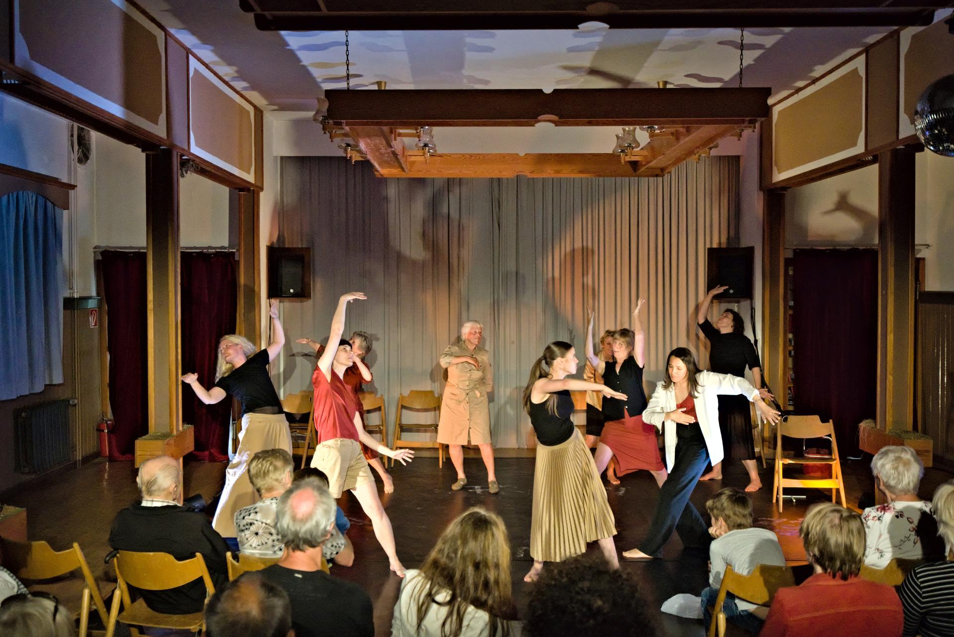 A group of nine dancers, from old to young, dancing on a stage