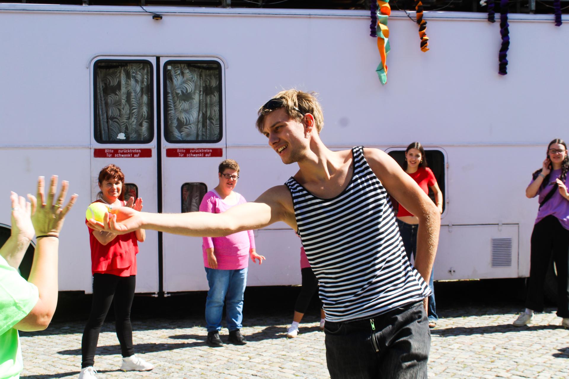 A dancer wearing a striped tank top is holding a ball in his outstretched hand. Several people are standing around him, clapping.