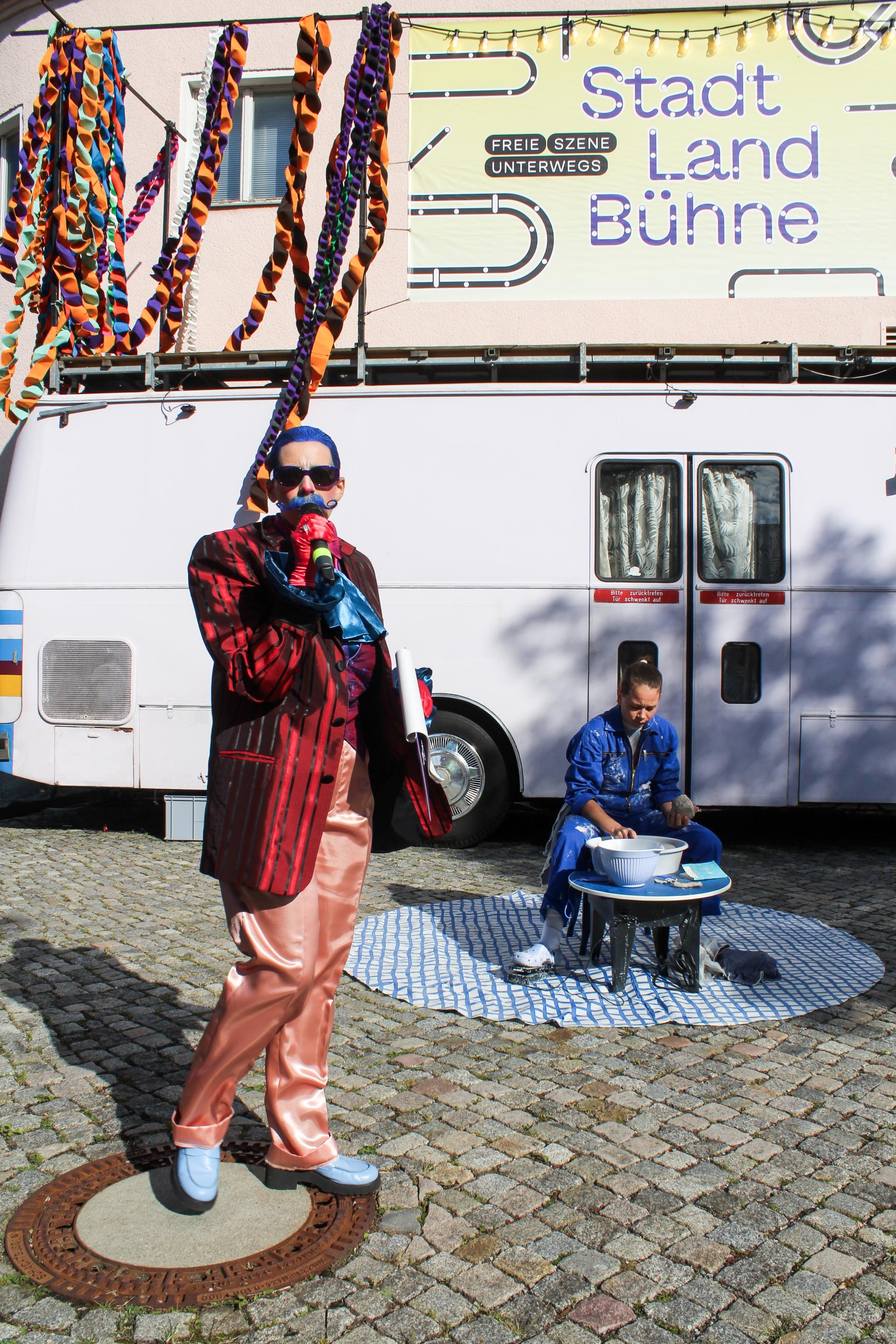 A host dressed in a colorful suit is speaking into a microphone. Behind her, a performer is sitting at a potter's wheel.