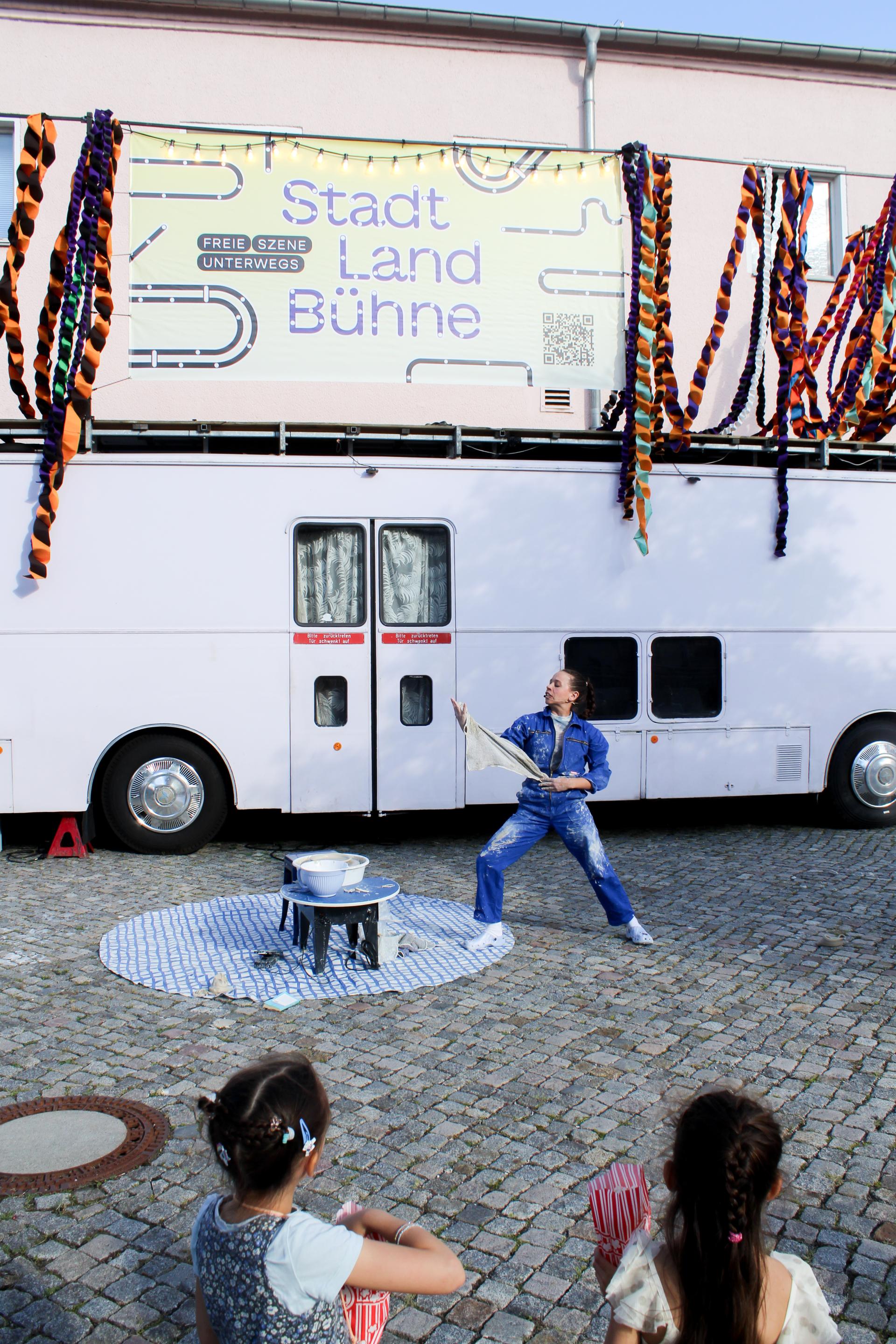 A performer in overalls stands next to a potter's wheel and speaks to the audience